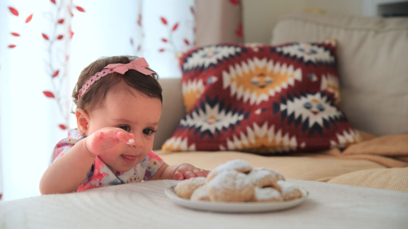 A little girl stands by a table reaching for a plate full of cookies.